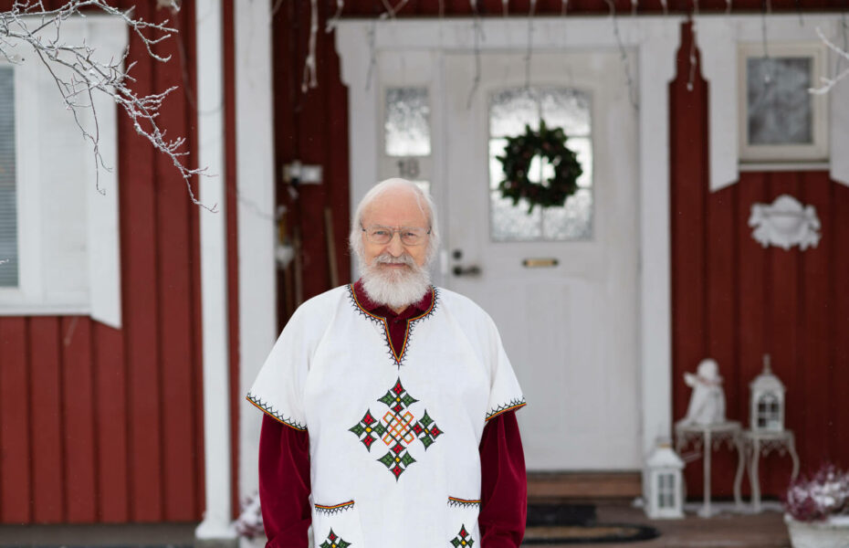 Seppo Väisänen seisoo omakotitalon ulko-oven edessä. Hänellä on valkoinen parta ja hiukset sekä silmälasit.