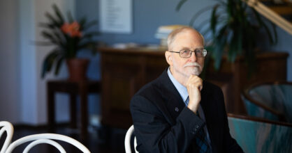 Mies, jolla on valkoinen parta nojaa käteensä sisällä ja istuu penkillä.