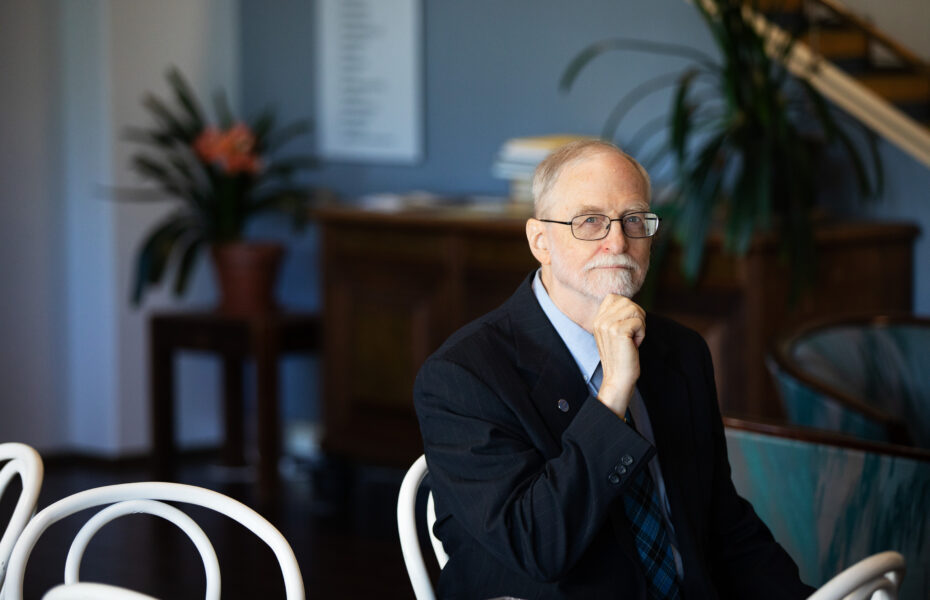 Mies, jolla on valkoinen parta nojaa käteensä sisällä ja istuu penkillä.