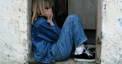 Tyttö istuu vasten seinää ja peittää kasvonsa käsillään.