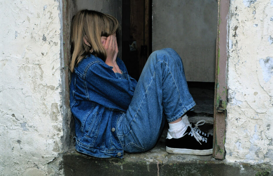 Tyttö istuu vasten seinää ja peittää kasvonsa käsillään.