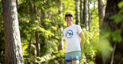 Susanna Ylinen seisoo metsässä ja hymyilee. Hänellä on lippalakki, t-paita ja shortsit.