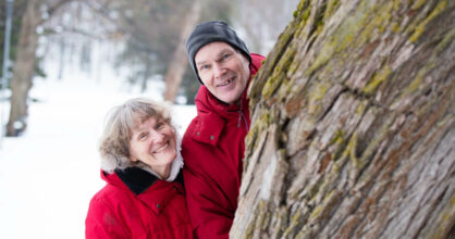 Susanne ja Kari Valkama kurkistavat paksun puun takaa lumisessa puistossa.