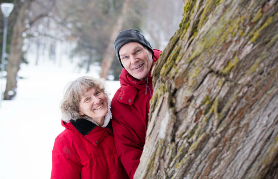 Susanne ja Kari Valkama kurkistavat paksun puun takaa lumisessa puistossa.