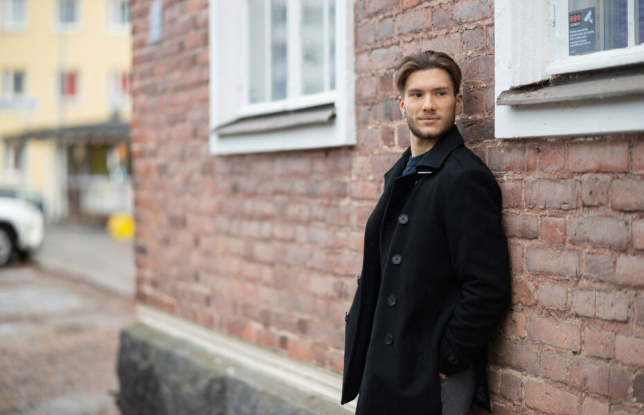 Mies nojaa tiilitalon seinään ulkona ja katsoo sivulle.