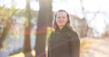 Pitkähiuksinen, keski-ikäinen nainen hymyilee. Taustalla on puita.