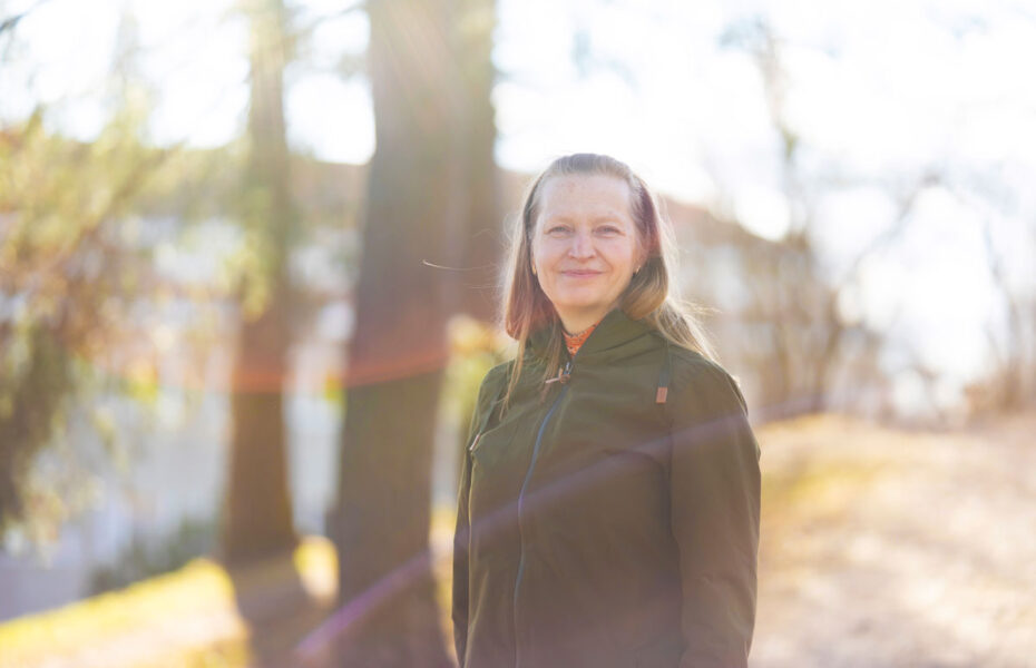 Pitkähiuksinen, keski-ikäinen nainen hymyilee. Taustalla on puita.