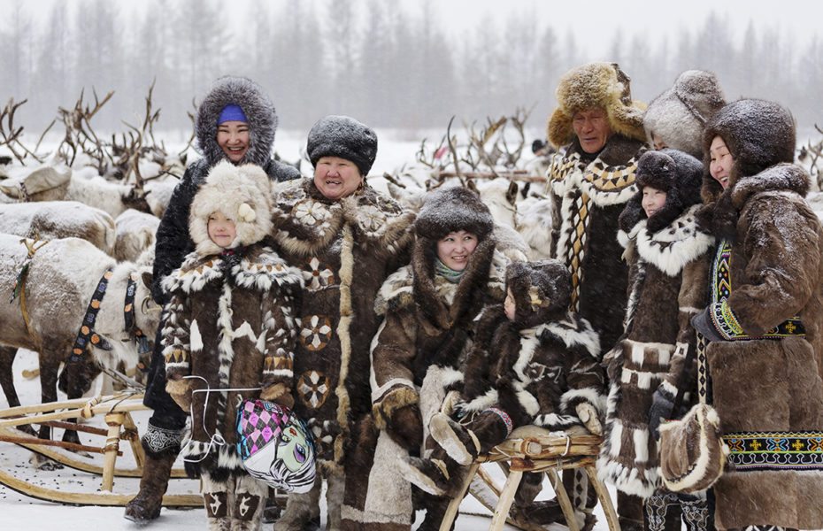 Ryhmä eri ikäisiä alkuperäiskansaan kuuluvia ihmisiä seisoo lumisateessa ja hymyilee. Heidän vaatteensa on tehty taljoista ja turkiksista, ja heidän takanaan on porolauma.