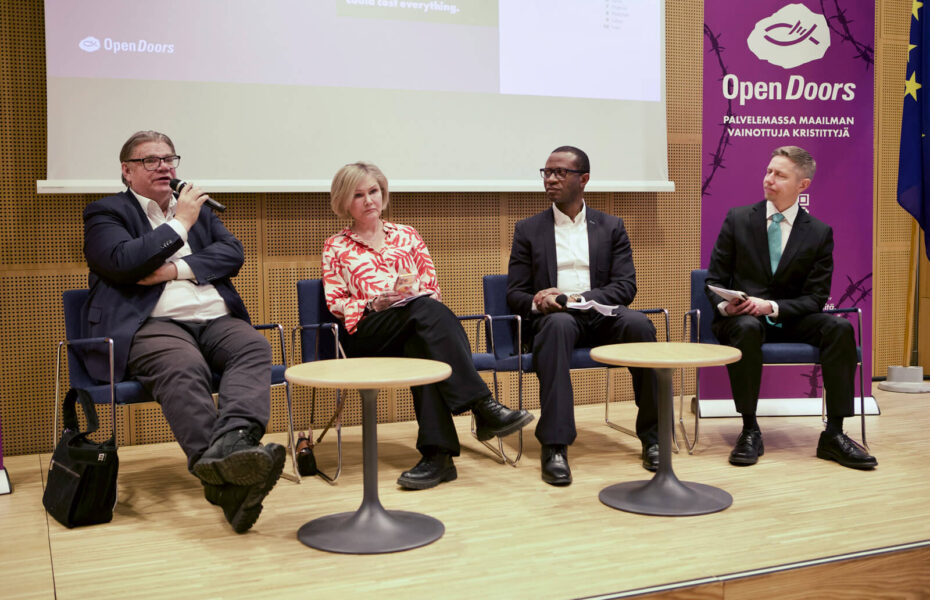 Neljä keskustelijaa istuu tuoleilla rivissä. Yksi heistä puhuu mikrofoniin.