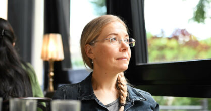 Ulla Barendsen-Riikonen istuu pöydän ääressä ja katsoo ikkunasta ulos. Hänellä on silmälasit, ja hänen hiuksensa on letitetty.