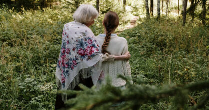 Vanha ja nuori nainen seisovat vierekkäin metsässä. Vanha nainen lohduttaa nuorempaa.