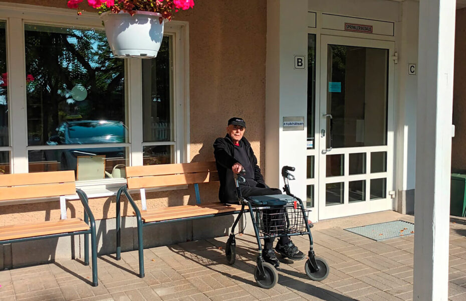 Iäkäs mies istuu penkillä. Hänen edessään on rollaattori.