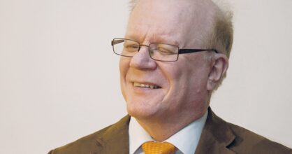 Vanhempi mieshenkilö hymyilee silmät kiinni. Silmälasit päässä ja ruskea puvuntakki kravatilla.