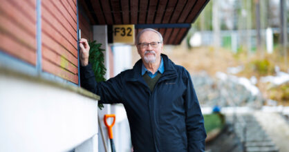 Mustaan takkiin pukeutunut harmaantunut ja silmälasipäinen mies nojaa seinään.