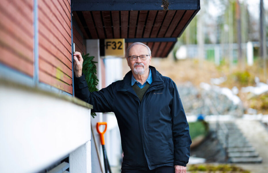 Mustaan takkiin pukeutunut harmaantunut ja silmälasipäinen mies nojaa seinään.